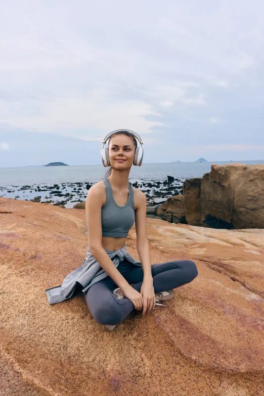 Young woman in activewear meditating on a rocky beach with headphones, promoting wellness and relaxation by the sea