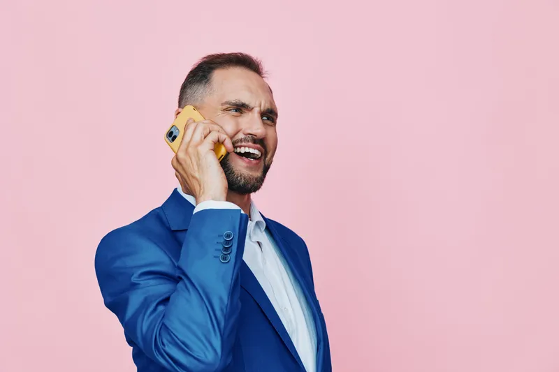 Professional man on a pink backdrop, wearing a blue suit, talking on a yellow phone, displaying confident energy and a keen business focus.