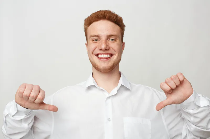 Smiling young man with red hair and beard wearing white shirt points thumbs at himself. Studio portrait on white background. Concept of confidence, self pride, and positive attitude.