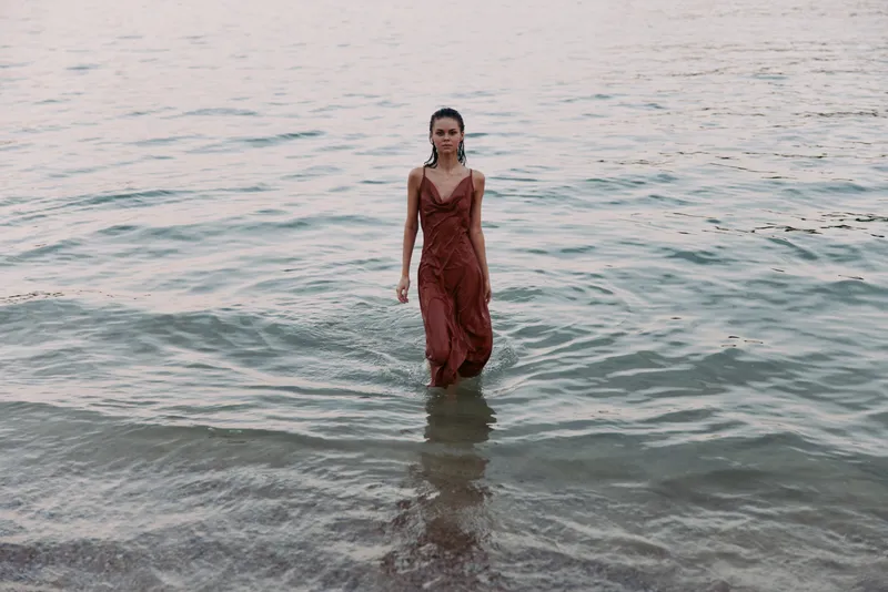 A solitary woman in a flowing red dress stands in shallow sea water near the shore, water laps at her ankles, creating a serene, graceful focal point against the waves.