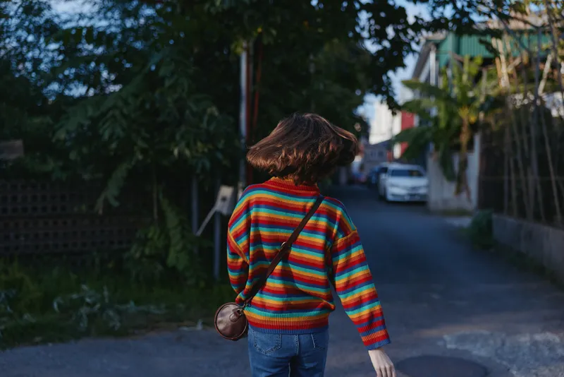 Striped sweater young woman walking suburban street with crossbody bag and bob haircut, mid-20s casual outfit and denim jeans, relaxed mood in evening light, lifestyle travel scene in quiet
