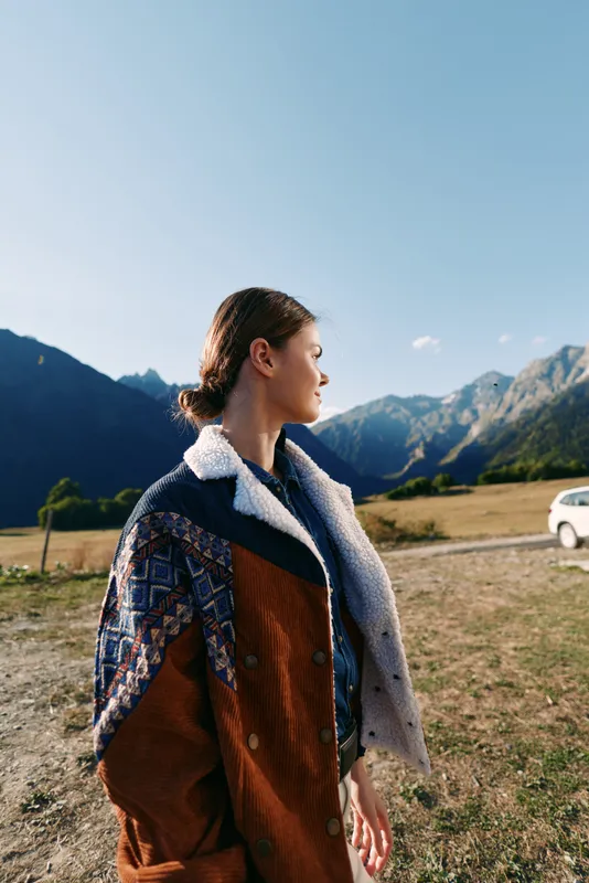 Woman in profile wearing a patterned coat stands in countryside landscape with mountains in the background and a parked car, autumn travel scene with warm natural light and calm mood.