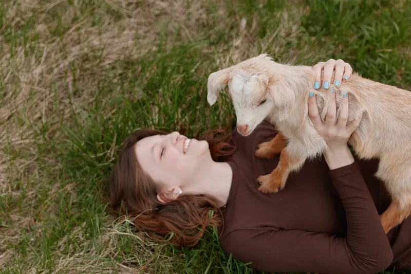 Young woman in a brown sweater lies on grass holding a small white baby goat while smiling and enjoying a peaceful moment outdoors in a natural lifestyle setting.