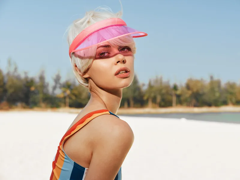 Portrait of a stylish young woman on a sunny beach, wearing a pink visor and colorful swimsuit, with bright light and shallow depth of field.