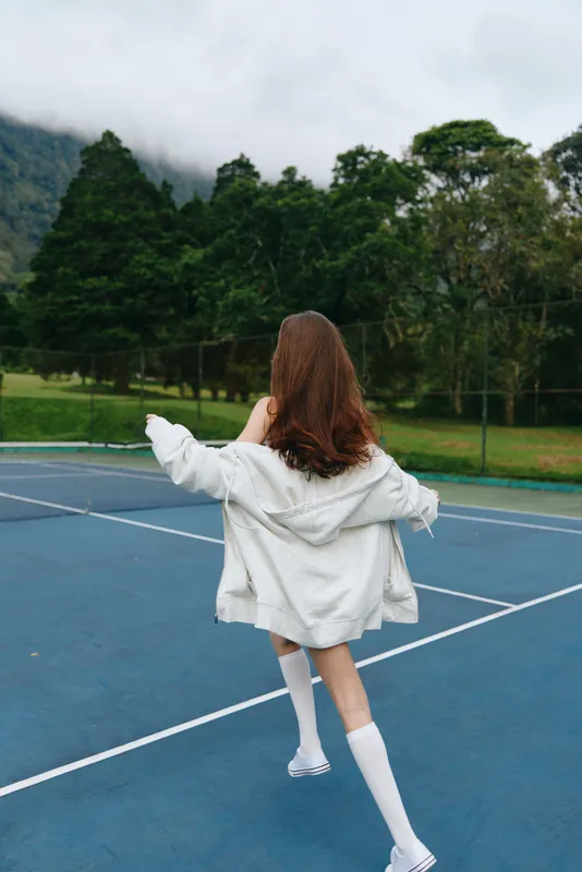 Young woman running on a tennis court, wearing a white hoodie and knee high socks, conveying joy and freedom in a natural green setting
