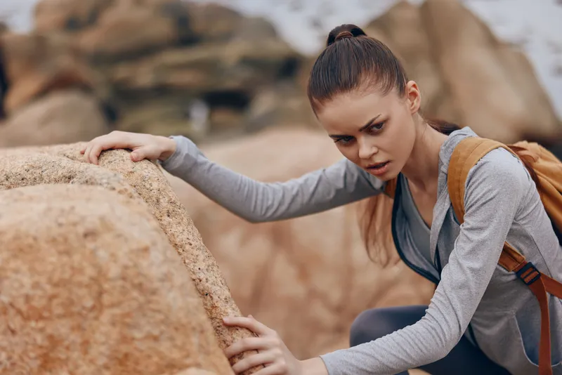Young woman climbing rocks at the beach, displaying determination and focus, with a casual athletic outfit in tones of gray, surrounded by natural scenery