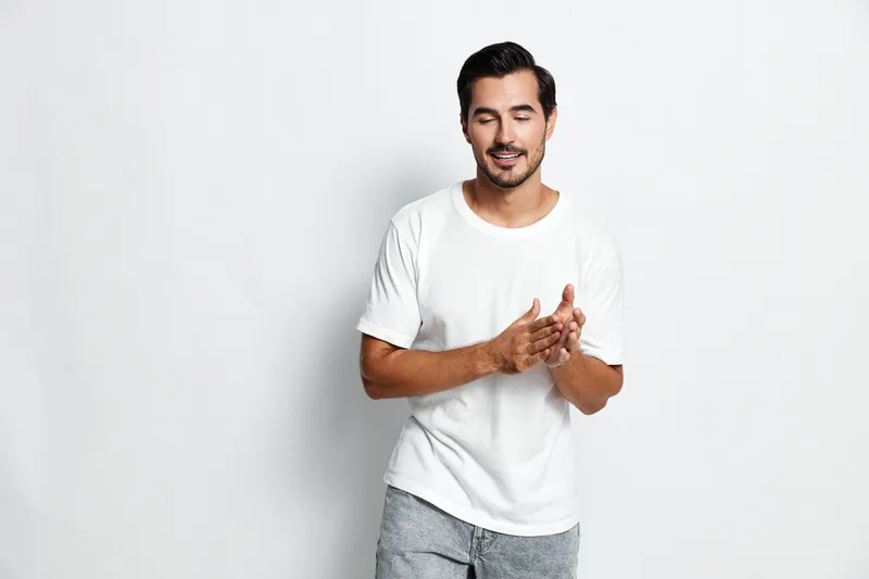 Casual smiling man wearing a white t-shirt gestures with hands while standing on a gray background. Positive vibe and friendly atmosphere captured in studio portrait.