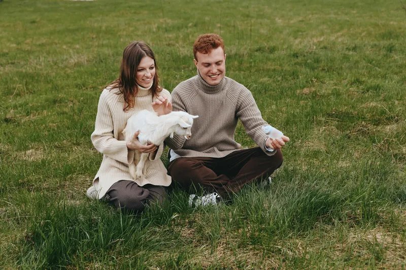 Couple sitting on grass with a playful goat kid, sharing a gentle moment outdoors. Cozy sweaters, natural light, warm countryside mood. They interact tenderly, suggesting lifestyle, wellness, and