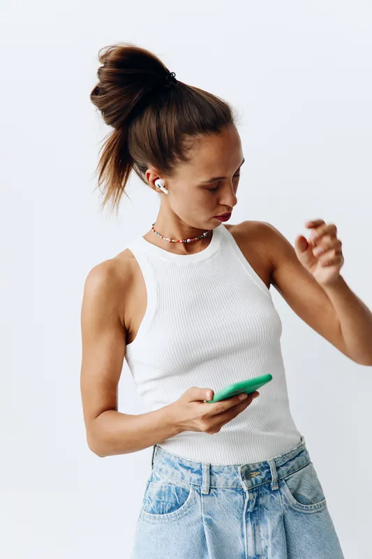 Молодая женщина поколения миллениалов слушает музыку через беспроводные наушники, подключенные к телефону, танцует и улыбается дома на белом фоне
