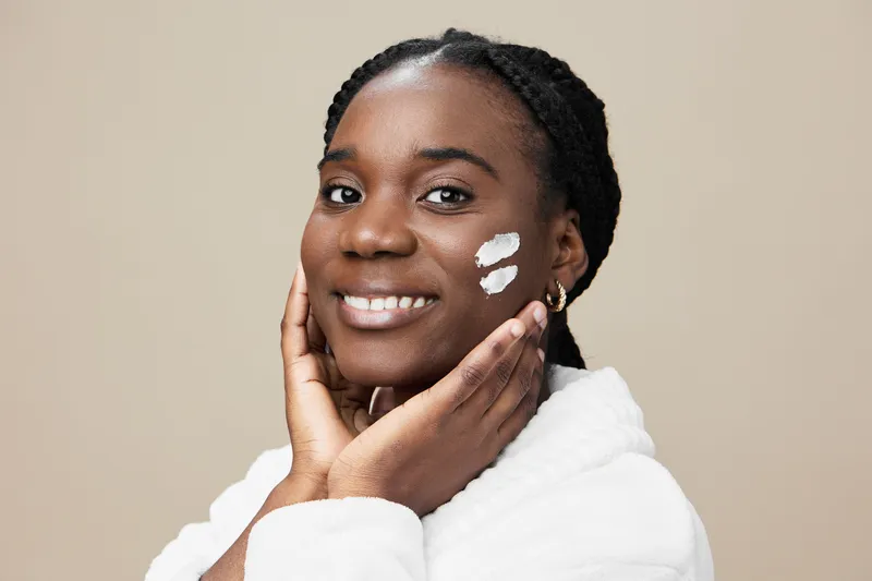 Smiling African woman applying skincare cream on her face, promoting healthy beauty routines with a warm, neutral background
