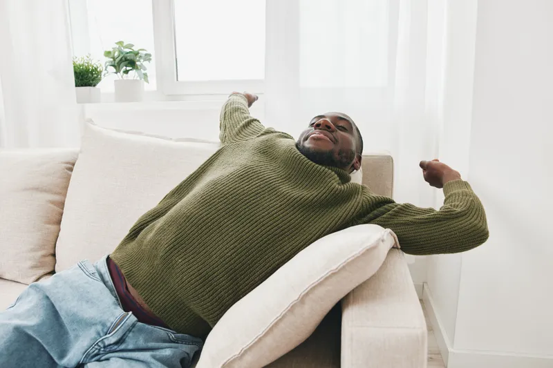 Relaxed young Black man in a cozy green sweater enjoying a peaceful moment while lounging on a beige sofa at home, exuding tranquility and comfort