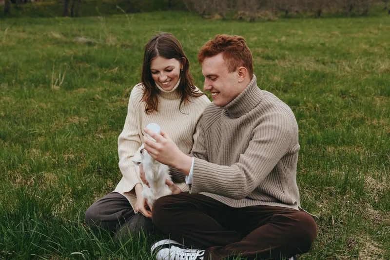 Young couple in cozy sweaters sits on grass enjoying playful moments with a small white baby goat in a natural outdoor setting during autumn.