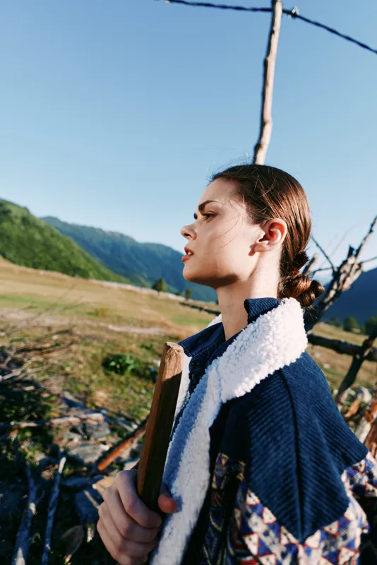 Woman portrait in countryside with mountains and field, rural profile of a young woman wearing a jacket and shearling, holding a wooden plank by a rustic fence in nature.