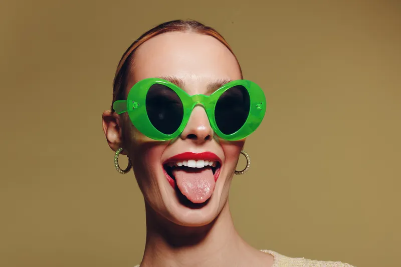 Young woman with green sunglasses sticks out tongue, smiling playfully. Studio portrait on beige background. Concept of fun, youth, fashion, and carefree attitude.