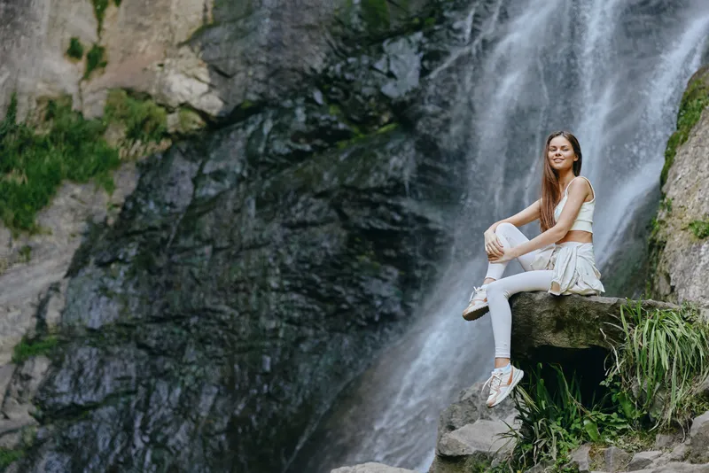 young woman sitting on rock near waterfall, wearing white activewear, surrounded by lush greenery and natural rocky landscape, enjoying outdoor adventure and nature.