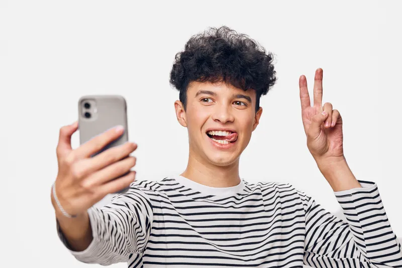 young man taking selfie with smartphone, making peace sign and funny face, wearing striped shirt, isolated on white background, casual style, cheerful mood
