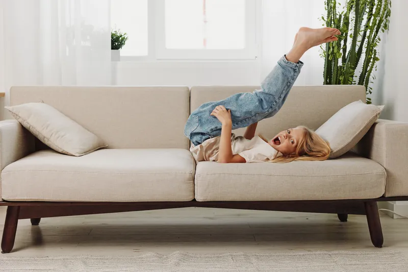 Happy child lying upside down on beige sofa in bright living room, playful mood, casual clothes, natural light, concept of childhood joy and home comfort.