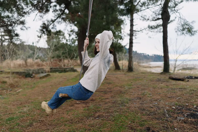 Person on a rope swing in a park, hoodie and jeans, enjoying outdoor nature by a lake with trees, breeze, and open sky, a carefree moment of leisure and fresh air.