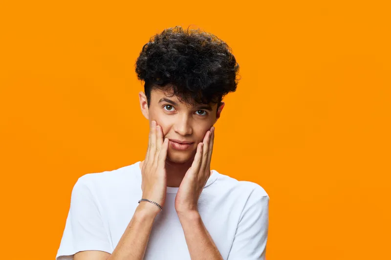 young man with curly hair touching face wearing casual white t-shirt on bright orange background, thoughtful expression, studio portrait