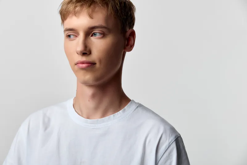 Young man wearing a white tee gazes to the left against a light studio backdrop, calm and thoughtful expression, clean casual style for fashion, lifestyle, or creative stock imagery.