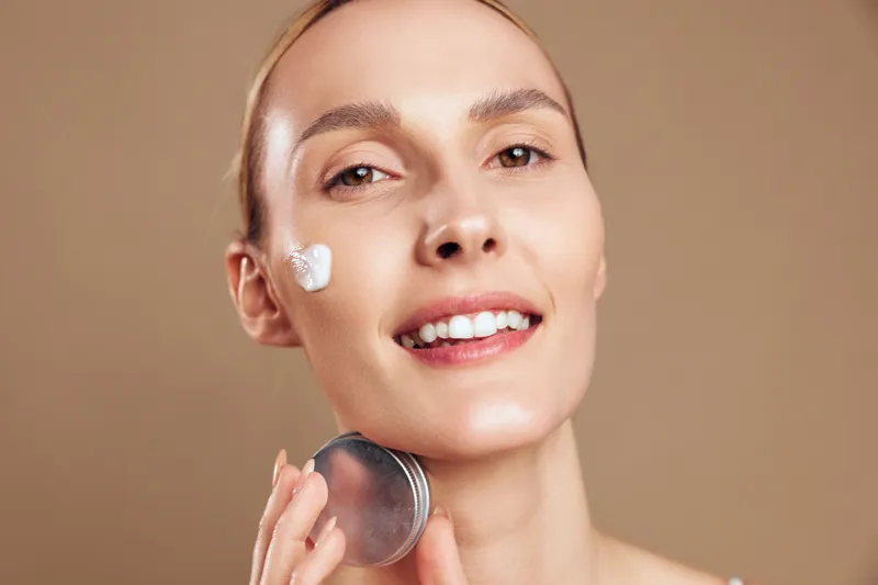 Woman in closeup performs skincare, applying cream to face, smiling with fresh glow, beauty spa routine, healthy smooth complexion, beige studio background, copy space, soft skin