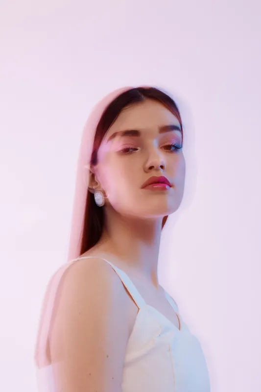 Young woman beauty fashion portrait with soft lighting on pink background. She wears white top and earrings, showcasing elegance and modern style. Concept of beauty and fashion.