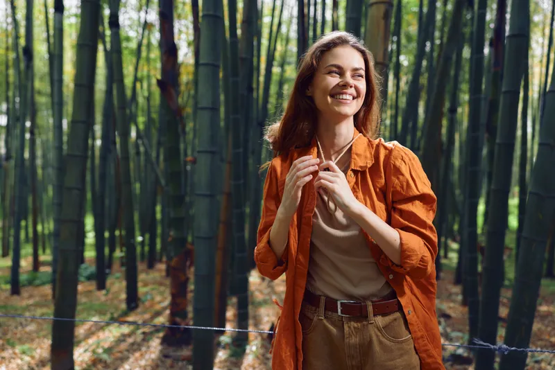 Woman smile in bamboo forest nature portrait, happy outdoors scene with sunlight and casual fashion jacket, walking among green trees and foliage for relaxation and scenic escape