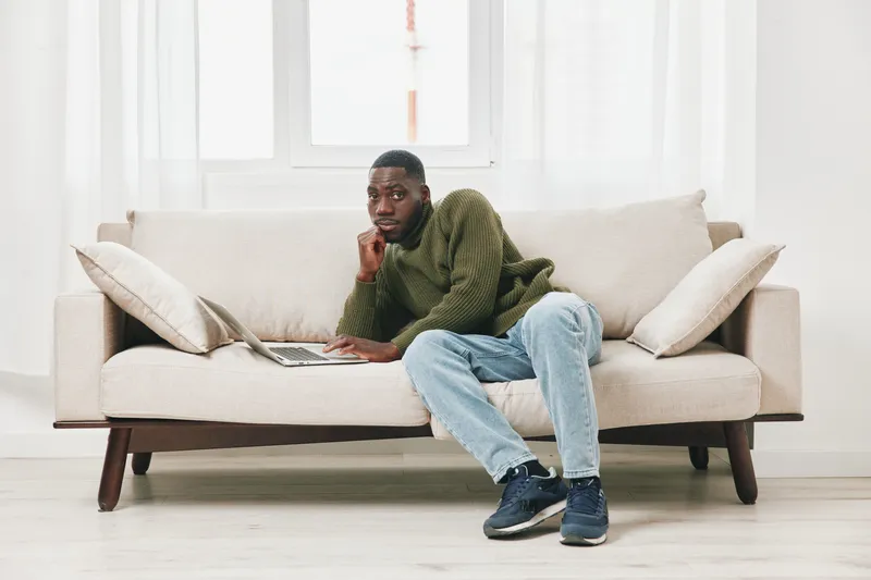 Thoughtful young Black man sitting on a modern couch while working on a laptop at home, dressed in a green sweater and jeans, conveying a sense of concentration and productivity