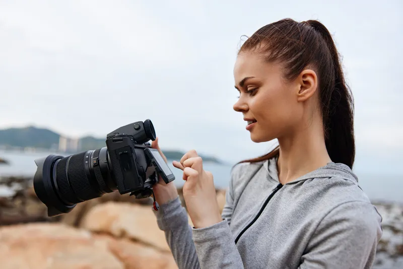 Young woman taking photos with a camera by the seaside, wearing a sports hoodie, with an engaged expression, capturing moments of nature and adventure