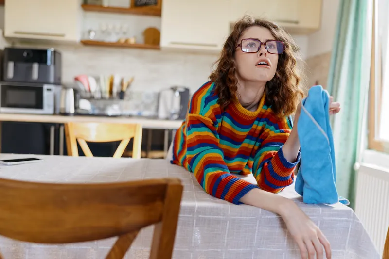 A woman with curly hair and glasses sits at a kitchen table in a bright striped sweater, holding a sock and looking up, capturing a candid home life moment of everyday tasks