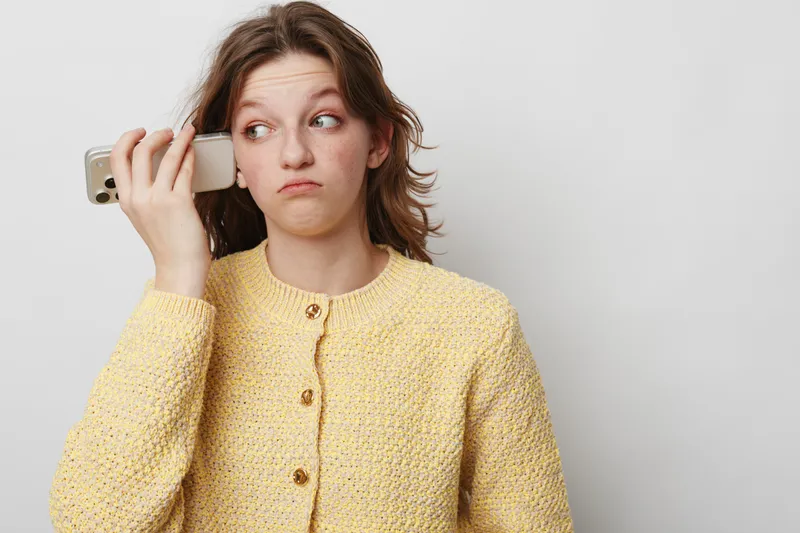 Young woman in yellow sweater holds smartphone to ear with skeptical expression. Isolated on white background. Concept of doubt, communication, and technology use in everyday life.