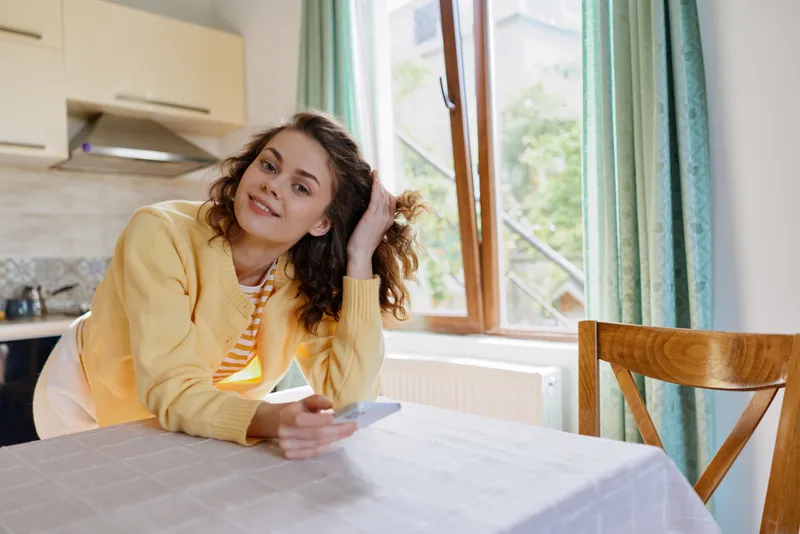 A young woman in a yellow cardigan smiles while resting her head on her hand at a sunlit dining table in a cozy home kitchen.