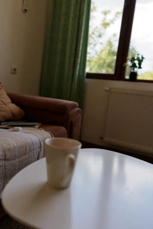 A white mug sits on a round coffee table in a cozy living room, with a brown sofa, green curtains and a sunlit window creating a calm morning atmosphere.