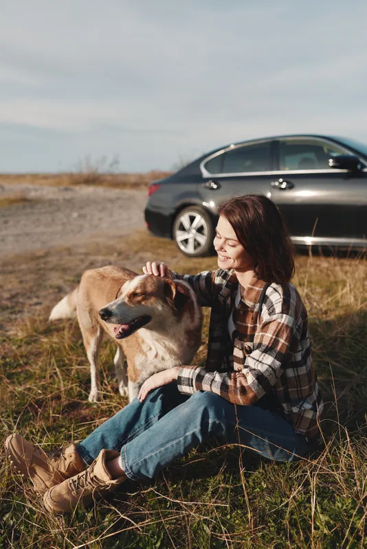 Woman sits on grass in a meadow with a dog beside her, car in the background, enjoying a peaceful outdoor moment and companionship on a sunny day.