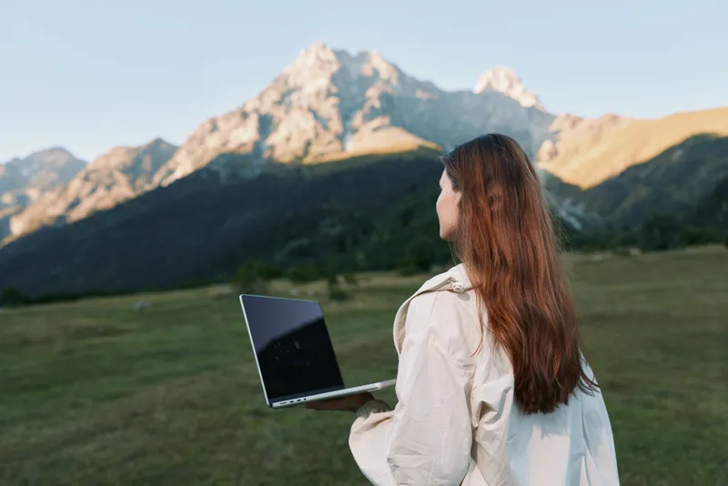 A woman stands in a grassy field with a laptop, facing distant mountains under clear skies. She wears light clothing and looks focused, conveying outdoor productivity, calm focus, and inventive work