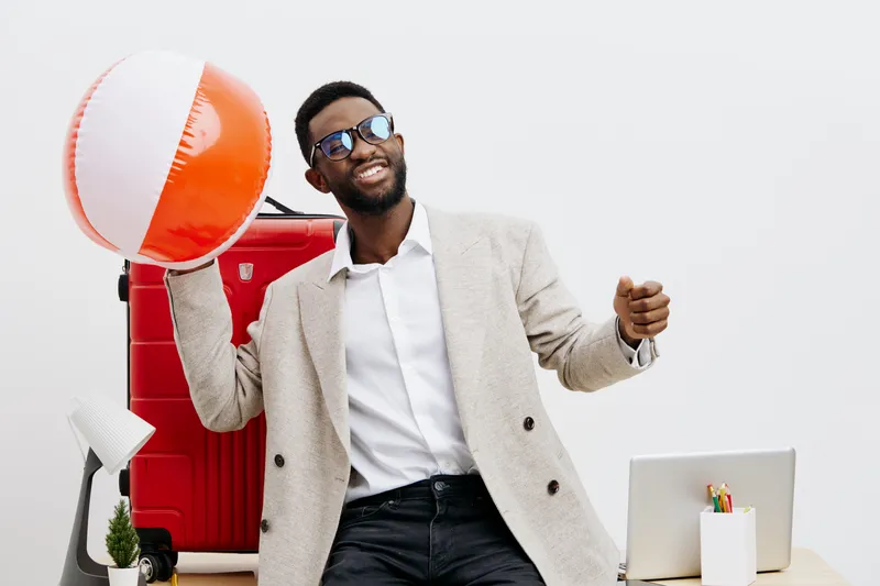 Cheerful smiling young African American man wearing sunglasses and casual beige blazer, holding a colorful beach ball over shoulder, standing in modern office environment with white background. People