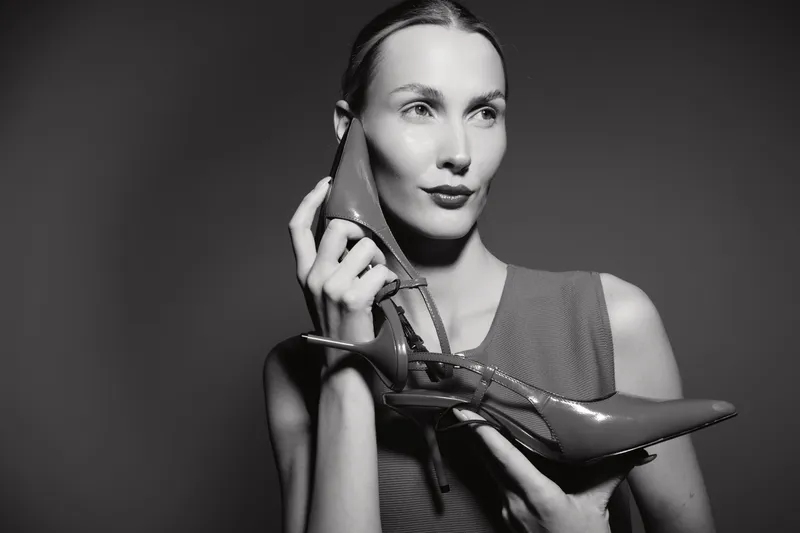 Caucasian woman with light skin holding shiny high heel shoe near face, wearing sleeveless top in studio. Concept for fashion, style, elegance, and beauty.
