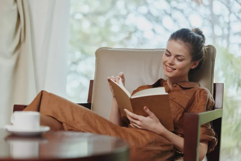 Молодая женщина в коричневом платье с радостью читает книгу, сидя в удобном кресле с чашкой кофе