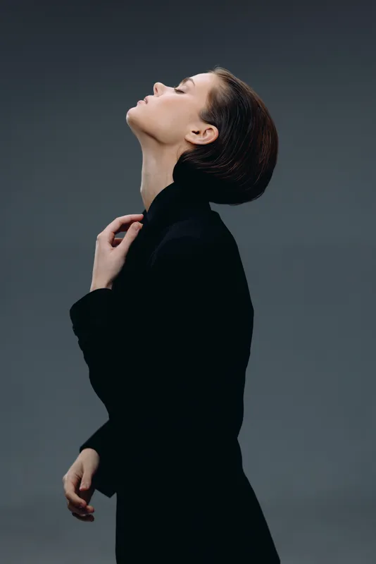 Elegant woman in black turtleneck posing with closed eyes against dark background, showcasing grace and calmness in a studio setting.