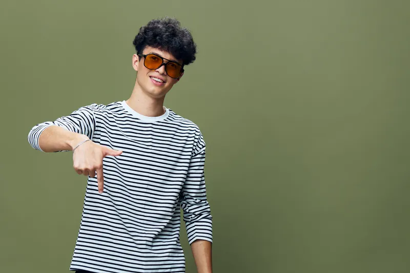 Young man wearing sunglasses and striped shirt pointing downwards with a confident smile against a green background in casual style.