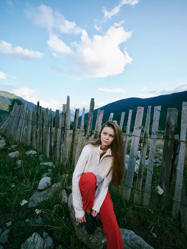 Woman in cozy sweater and red pants sits by a rustic fence in a green hillside landscape, capturing a quiet countryside mood for lifestyle, travel and outdoor photography