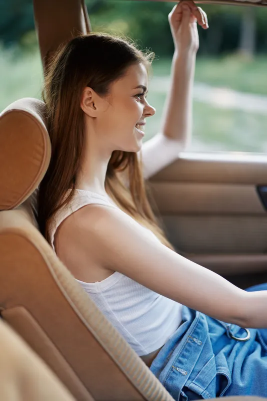 Young woman smiling in a car, wearing casual outfit, enjoying a sunny day, with green background and relaxed vibe. KEY: woman, smile, car, casual, sunlight, happiness, relaxation, summer, joy, beauty