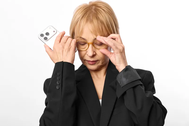 Senior woman adjusting eyeglasses while holding a smartphone, portrait on studio white background, showing headache, stress, eyes strain, health communication