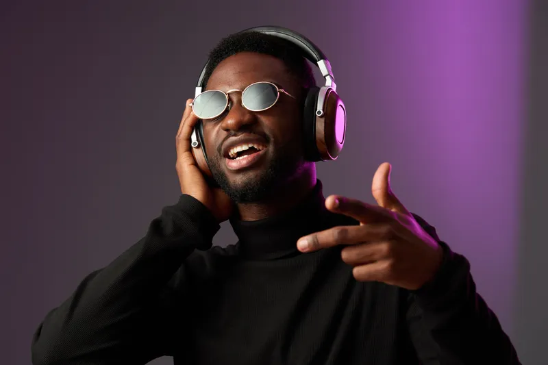 Young Black man enjoying music with headphones, wearing sunglasses against a purple and gray background, expressing joy and confidence in a modern style