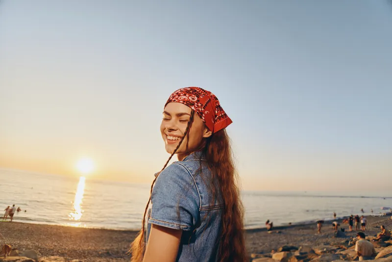 Smiling woman in modern boho-western denim street style enjoying sunset at beach with film color effect, creating warm and relaxed lifestyle atmosphere.
