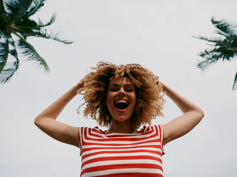 Joyful woman with curly hair in a red and white striped shirt smiles broadly under a bright sky, arms raised, palm trees framing a carefree outdoor moment.