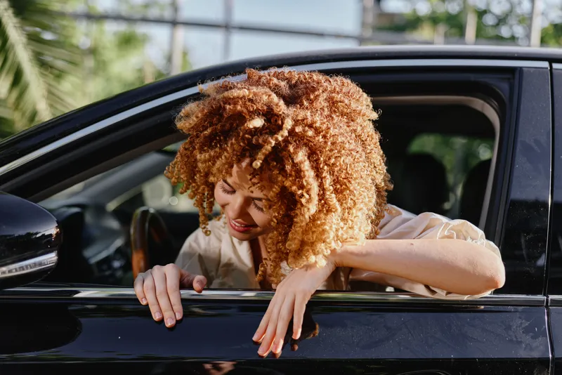 woman with curly hair leaning out car window smiling with sunlight and green trees background, casual clothing, relaxed moment, natural light
