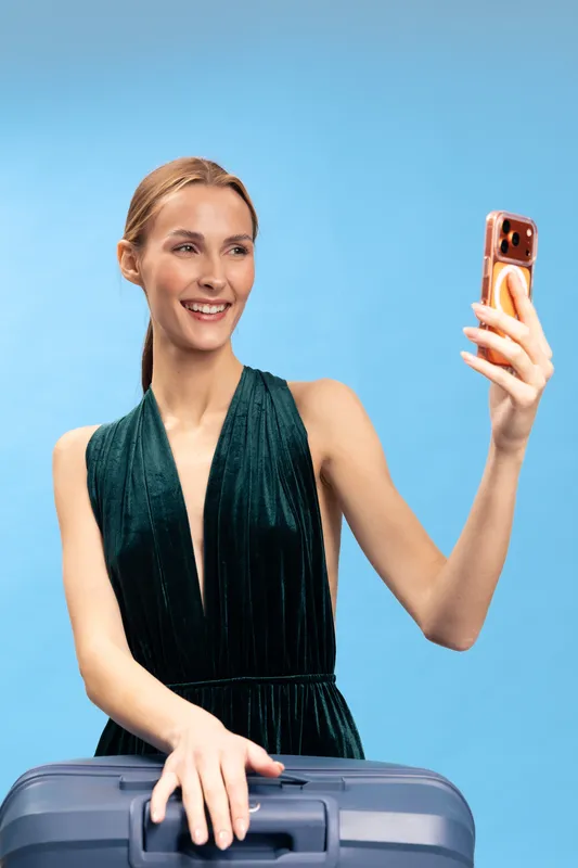 Young woman in green dress takes selfie with smartphone while holding suitcase on blue background. Concept of travel, vacation, and modern lifestyle with technology and happiness.