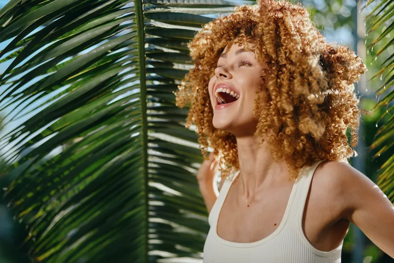 laughing woman with curly hair enjoying tropical sunlight near large green palm leaves outdoors in bright summer day feeling joyful and carefree
