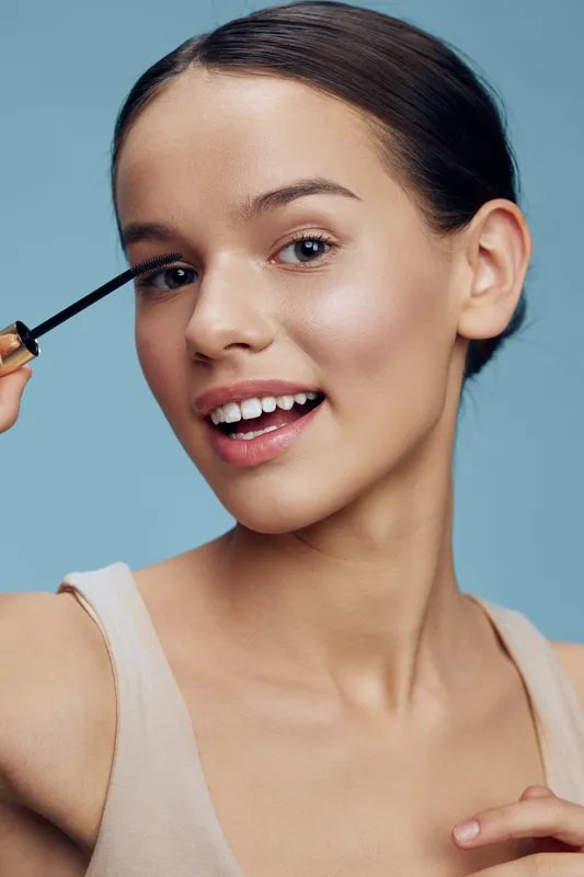 Young woman applying mascara on her eyelashes, showcasing natural beauty with a smile, against a soft blue background